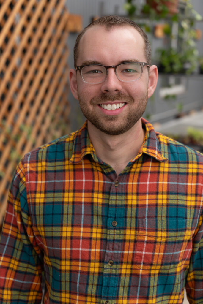 Brandon wears a colorful flannel for a headshot in a greenhouse with a trellis in the background.
